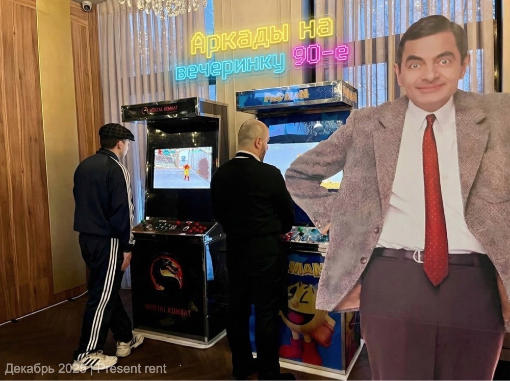 Два аркадных автомата в фойе вечеринки в стиле 90-х, один с Mortal Kombat, другой с Pac-Man, а также картонная фигура персонажа с улыбкой.