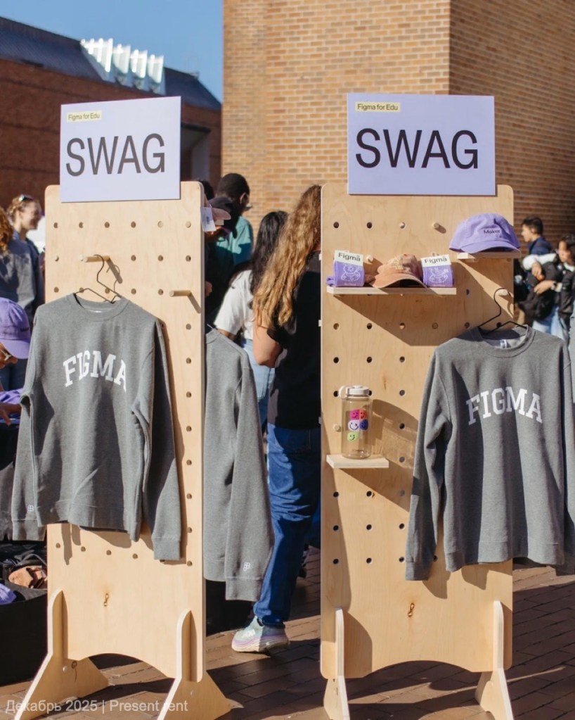 Стенд из фанеры с вешалками и полками для мерча Figma, на котором выставлены свитшоты и кепки. Сзади видны люди, присутствующие на мероприятии.