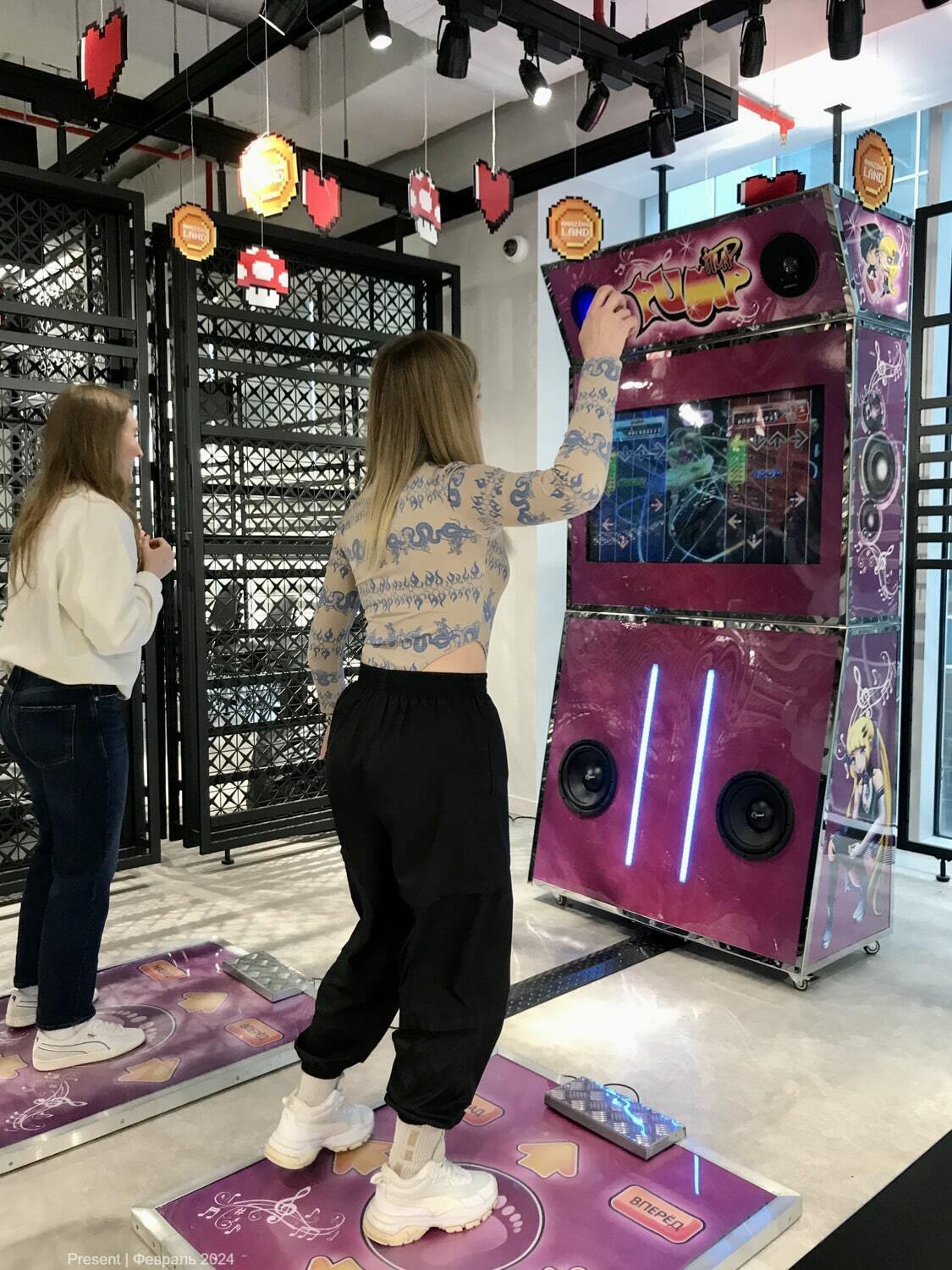 Девушка танцует на игровом автомате DDR в офисе. На экране автомата отображаются стрелки, соответствующие музыкальному ритму. Вокруг неё висят яркие декорации в виде пиксельных сердечек и грибов как в аркадном мире