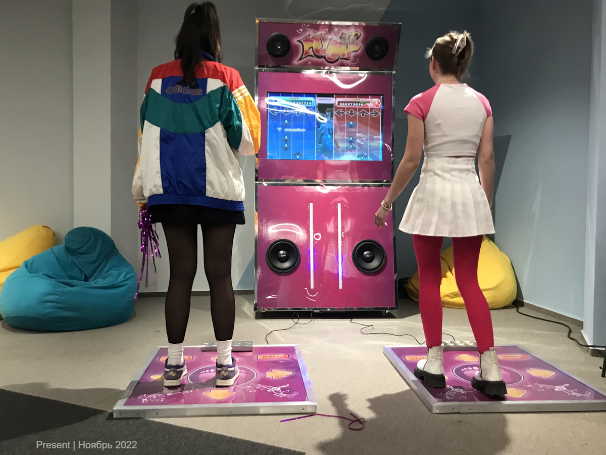 Две девушки участвуют в танцевальном батле на игровом автомате в офисе. Одна из них одета в разноцветную куртку и черную юбку, другая в костюм с розовыми леггинсами и белой юбкой. Вокруг расположены уютные сиденья- мешки. На экране автомата отображаются стрелки для танца.