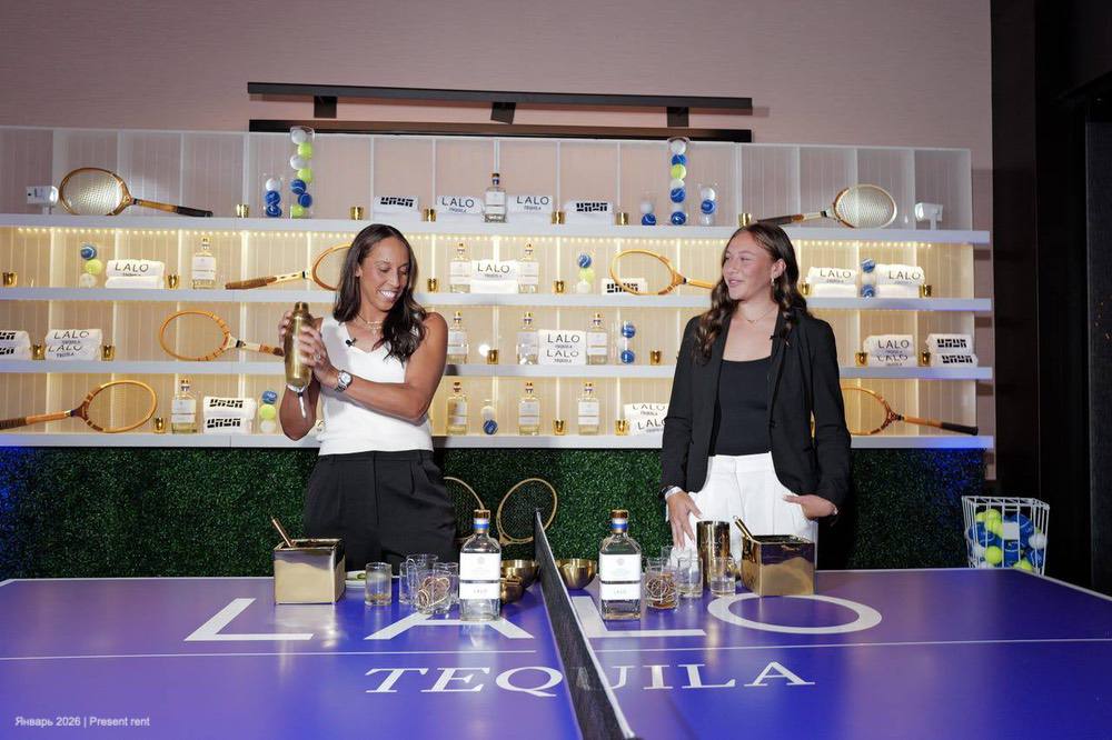 Две женщины стоят за столом с напитками и занимаются коктейльной подготовкой на фоне стенда с бутылками текилы и теннисными ракетками.