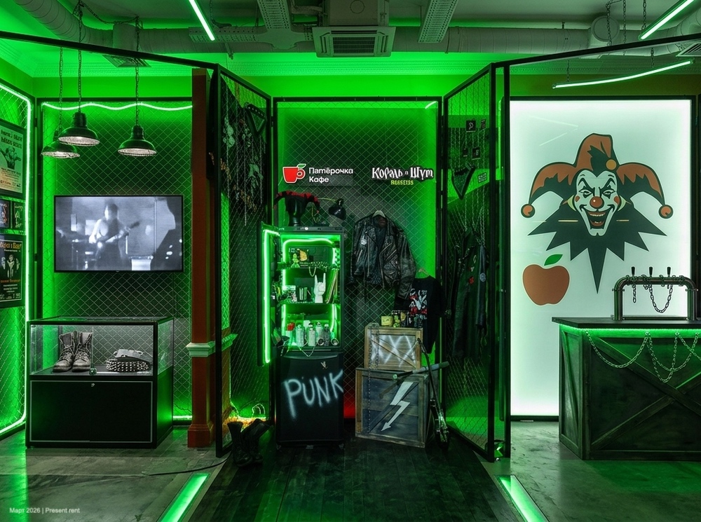 Интерьер кафе с темной темой и неоновым зеленым освещением, показывающий стиль рок-панк. На стенах висят кожаные куртки и различные аксессуары, а также видно черно-белое видео. Стойка оформлена с изображением шута и надписью 'PUNK'.