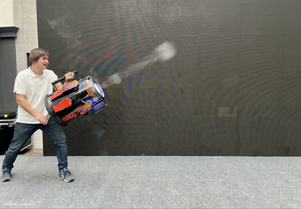 Молодой человек в белой рубашке держит в руках яркую и большую игрушечную пушку с эффектом выстрела на фоне черной стены.