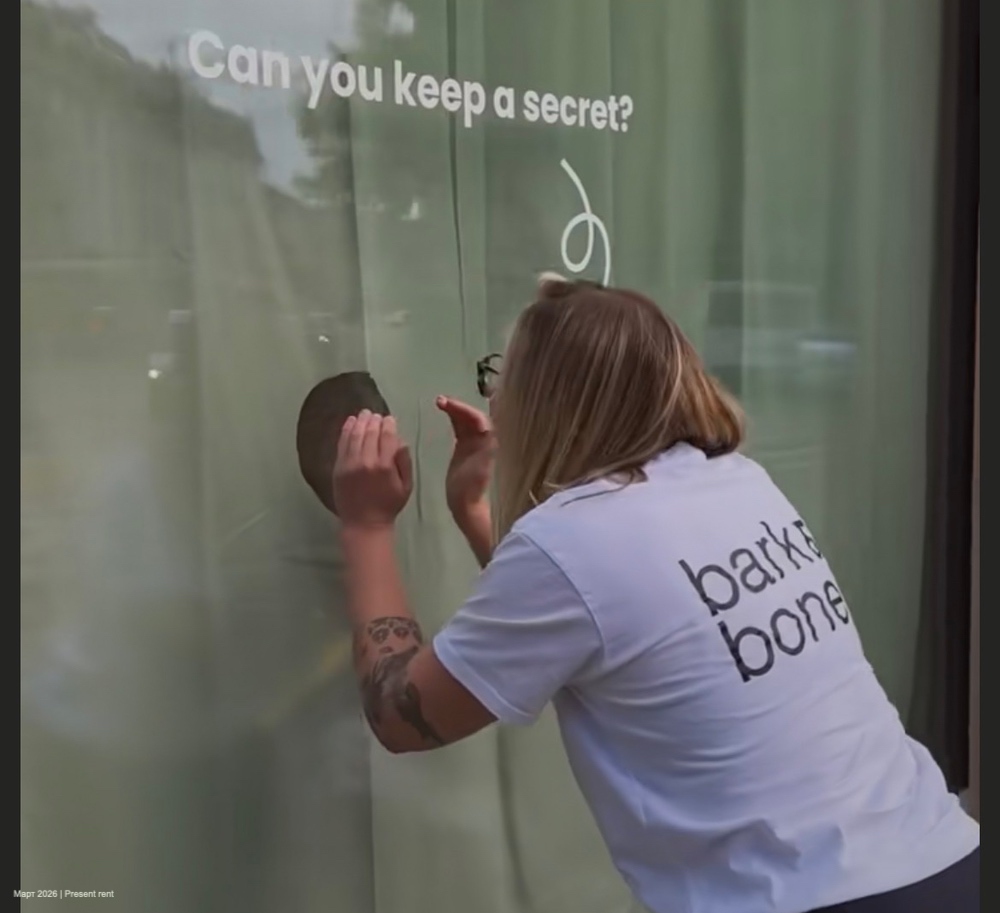 Женщина в белой футболке с надписью 'bark and bone' приклоняется к окну с прозрачной занавеской, пытаясь рассмотреть что-то внутри. На окне написано 'Can you keep a secret?'
