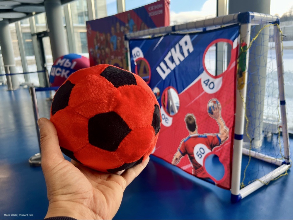 Человек держит красный и черный пушистый футбольный мяч на фоне спортивного стенда с целями и изображением игрока.